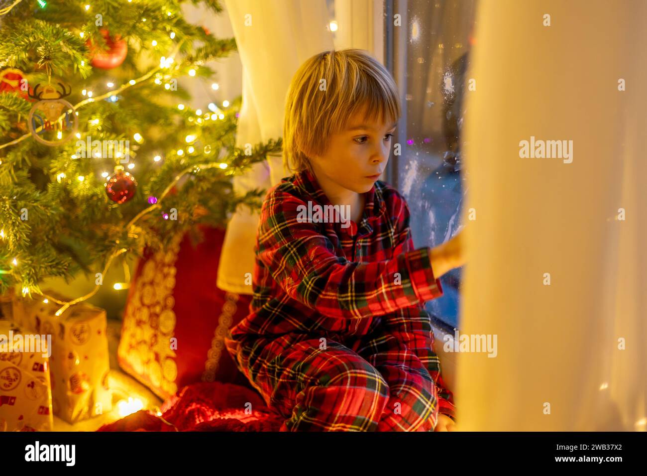 Niedliches Kind, das am Fenster sitzt, draußen nach dem Weihnachtsmann sucht und Kekse isst Stockfoto