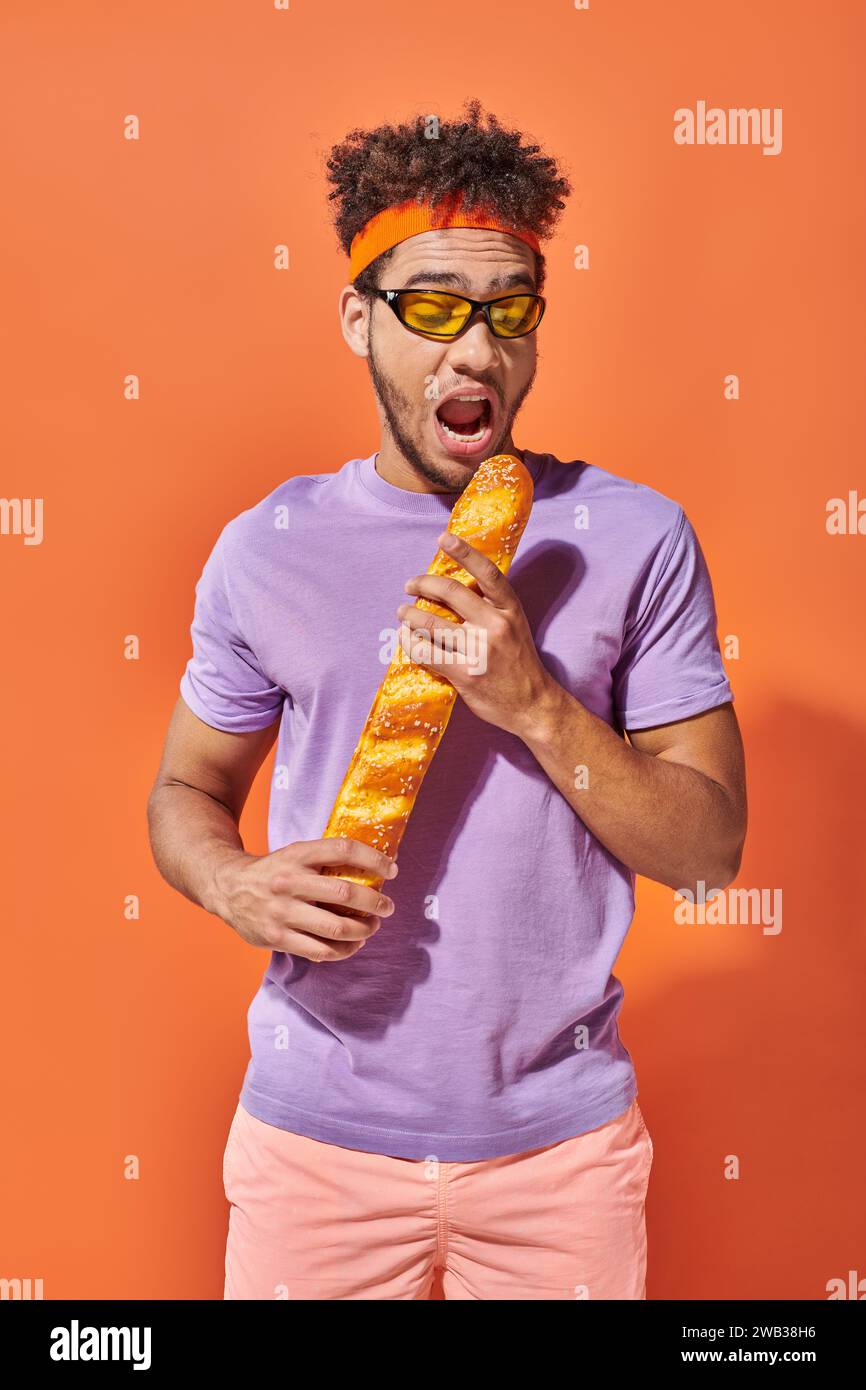 Junger afroamerikanischer Mann in Sonnenbrille und Stirnband, der frisches Baguette auf orangem Hintergrund beißt Stockfoto