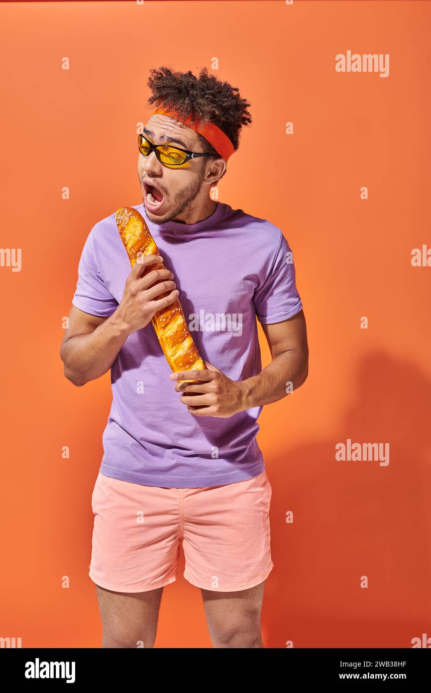 Lustiger afroamerikanischer Mann in Sonnenbrille und Haarreif, der frisches Baguette auf orangem Hintergrund beißt Stockfoto