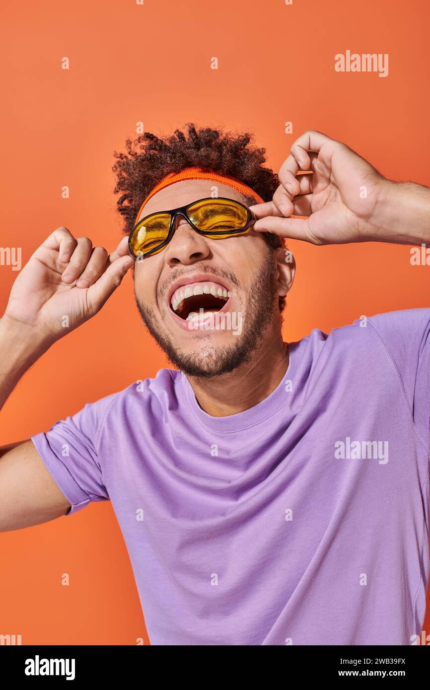 Glücklicher afroamerikanischer Mann mit Haarreif lächelnd und Sonnenbrille auf orangefarbenem Hintergrund Stockfoto