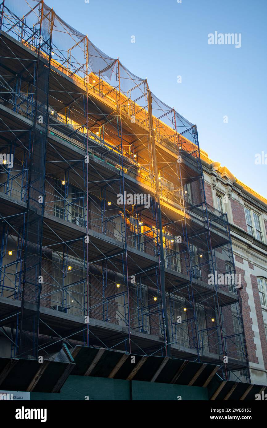 Die Silhouette eines Stadtgebäudes wird vom warmen, orangen Licht der untergehenden Sonne beleuchtet, während ein Gerüstnetz das Gebäude umgibt Stockfoto