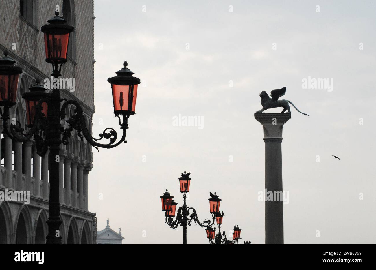 Eine wunderbare perspektivische Aussicht auf eine Reihe von Leuchtenpfosten mit roten Tönen auf dem Markusplatz von Venedig, Italien. Beachten Sie die Seegülle im Flug. Stockfoto