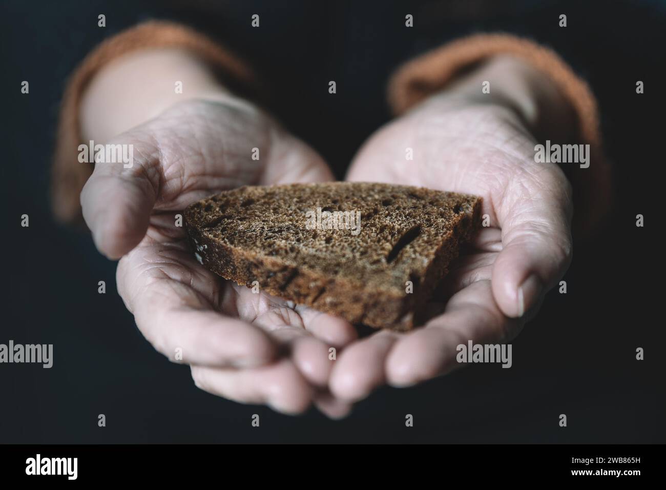 Eine alte Rentnerin hält ein Stück Schwarzbrot auf dunklem Hintergrund in der Hand. Das Konzept von Hunger und Armut. Stockfoto