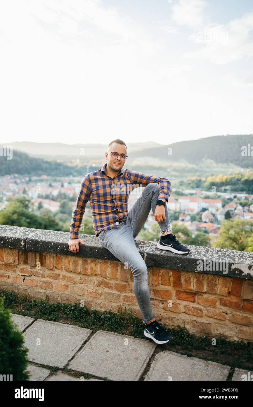 Ein junger Mann im Hemd sitzt während einer Reise lächelnd auf einer Burgmauer Stockfoto