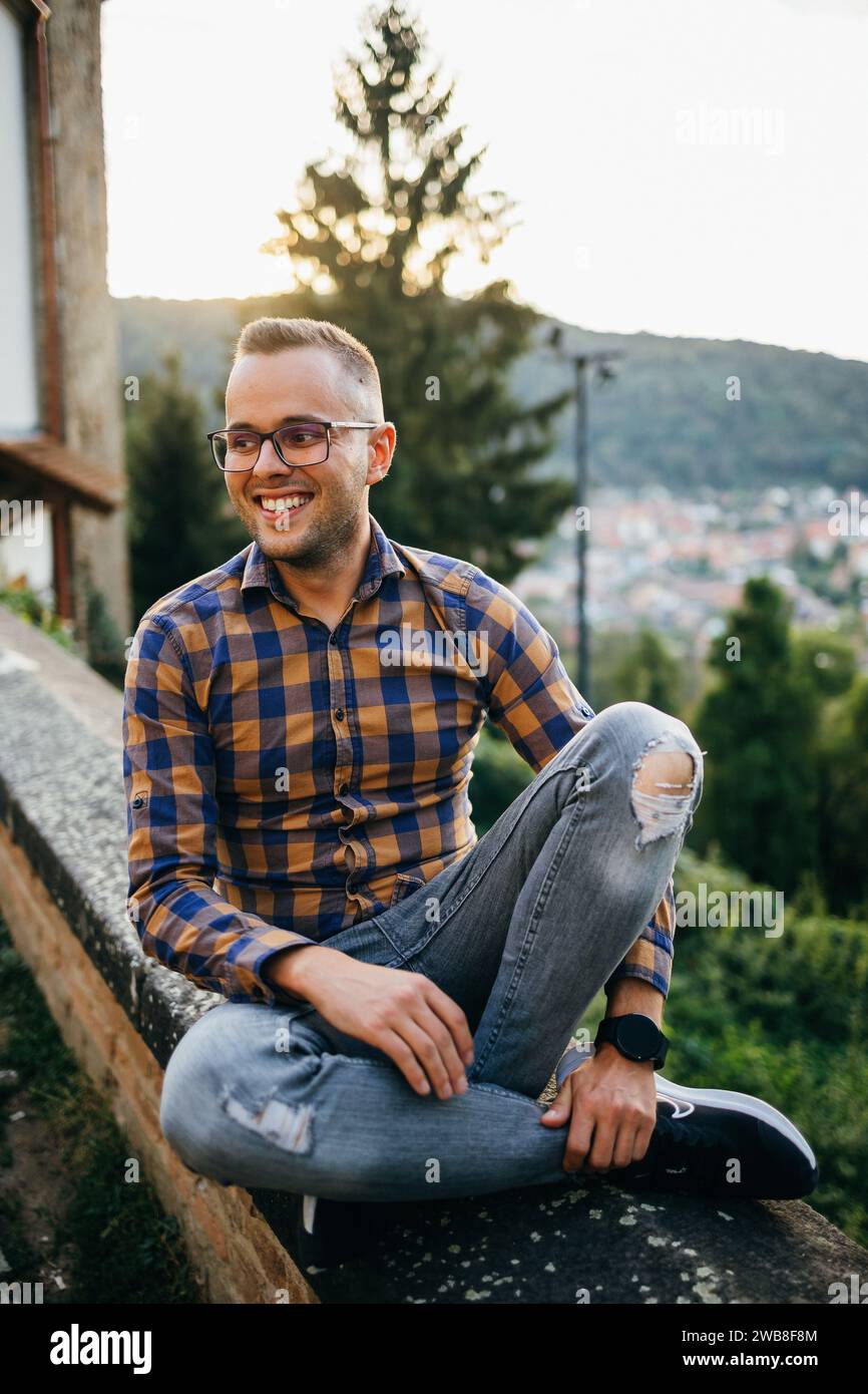 Ein junger Mann im Hemd sitzt lächelnd an einer Burgmauer Stockfoto