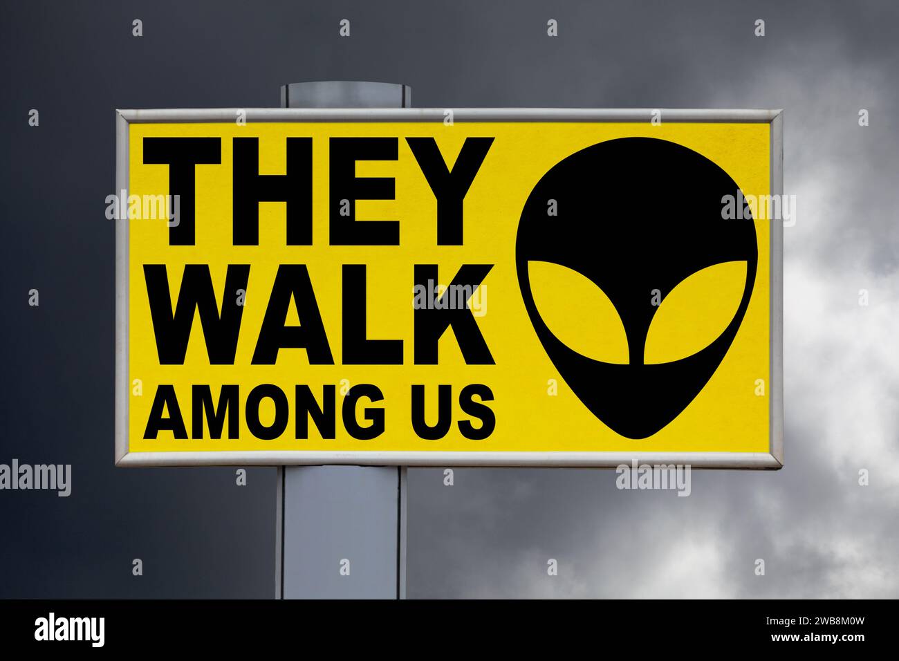 Nahaufnahme auf einer gelben Plakatwand vor einem bewölkten Himmel mit der Nachricht „They Walk Amony US“ in der Mitte neben einem Alien-Gesicht. Stockfoto