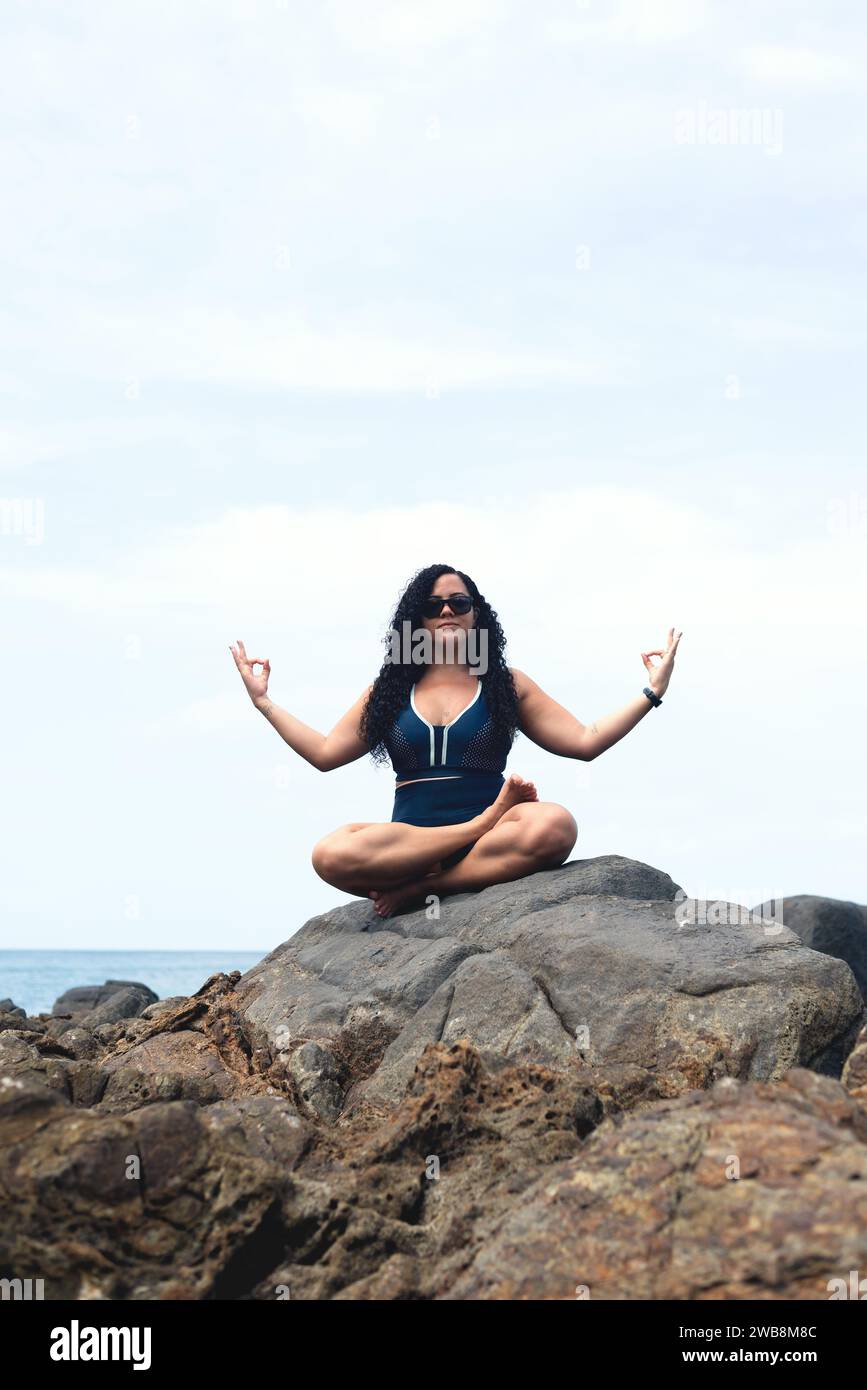 Schöne, lockige Frau, die auf dem Felsen eines Strandes sitzt und Yoga macht. Glückliche Person auf Reisen. Stockfoto