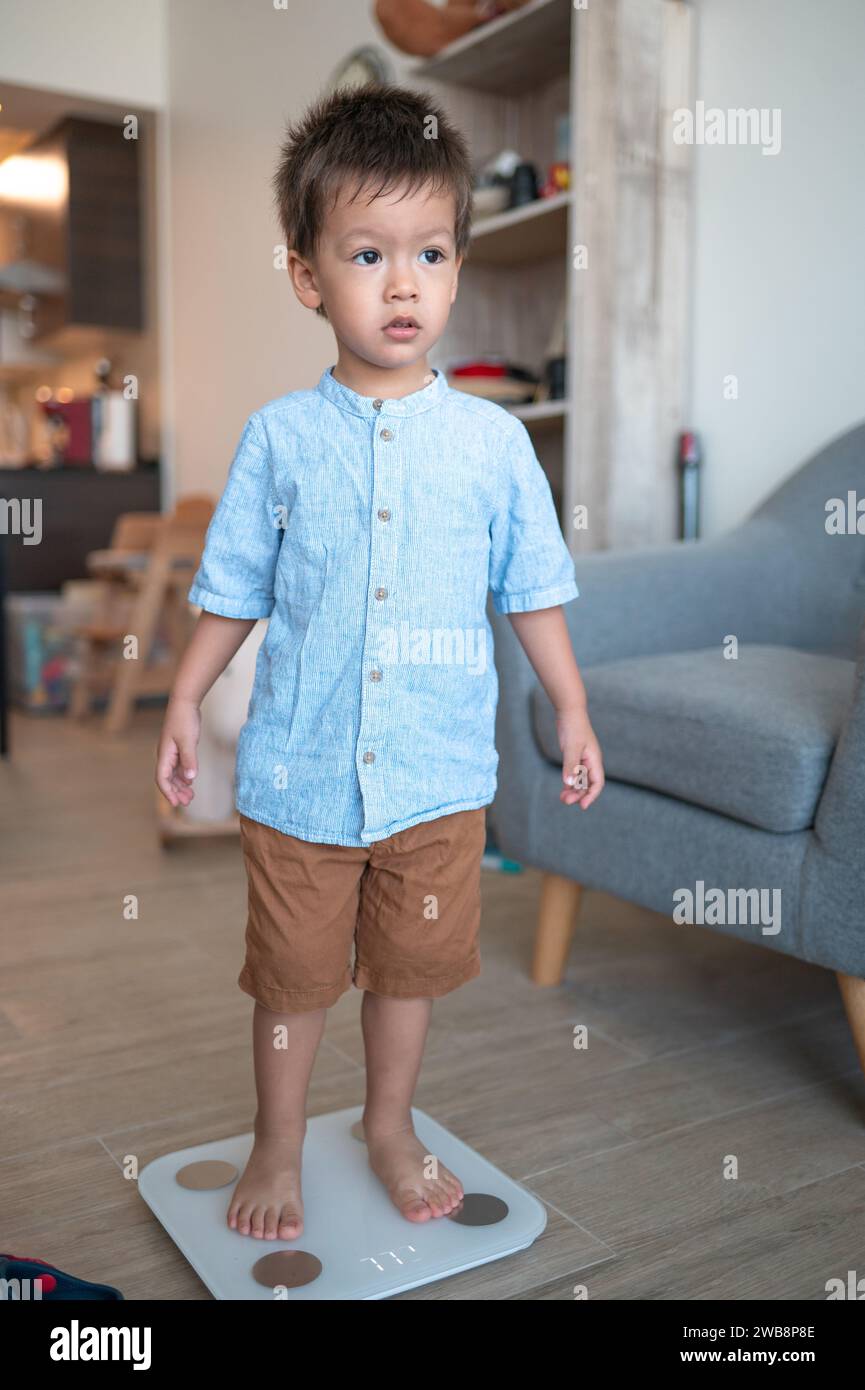 Zweieinhalb Jahre alter Junge, gekleidet in einem Hemd und bermuda-Shorts, der einen Schritt hin zu gesunden Gewohnheiten macht. Barfuß auf einer digitalen Smart Scale i stehen Stockfoto
