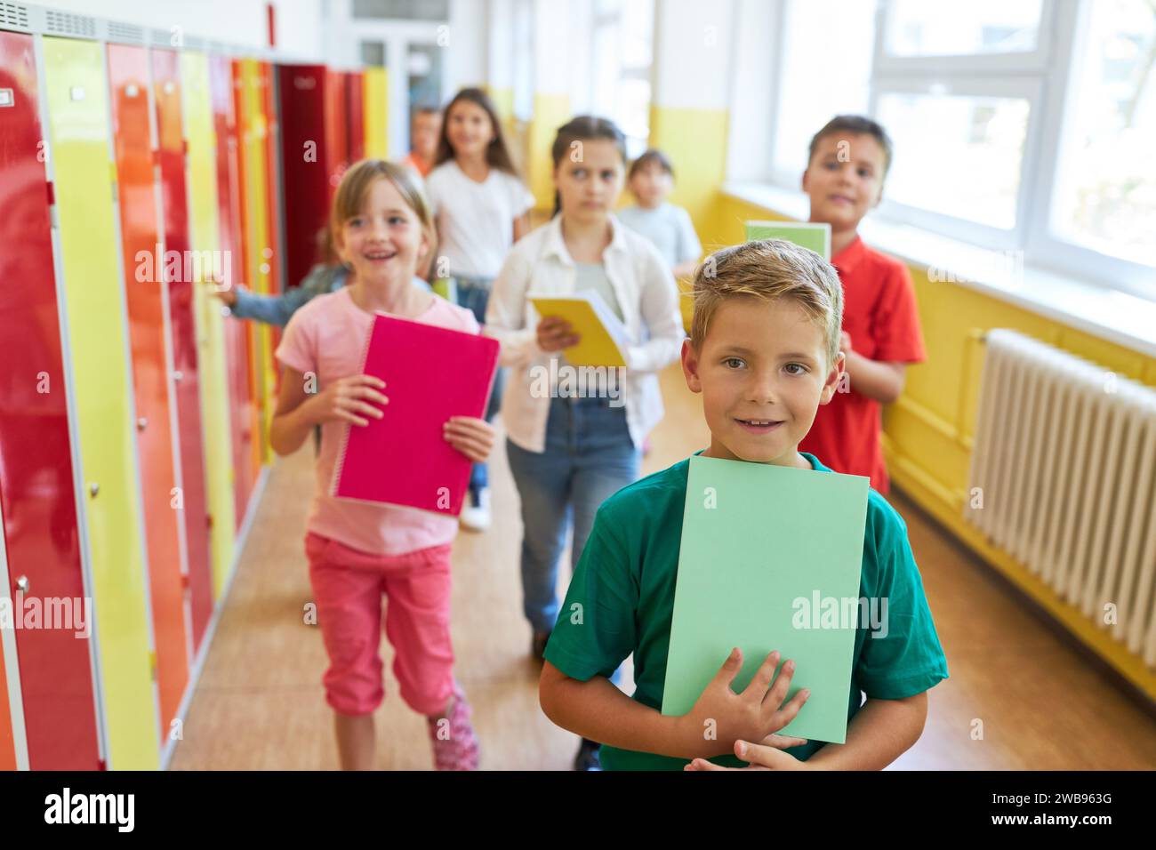 Junge, der ein Buch hält, während er mit Klassenkameraden in der Nähe von Schließfächern in der Schule spaziert Stockfoto