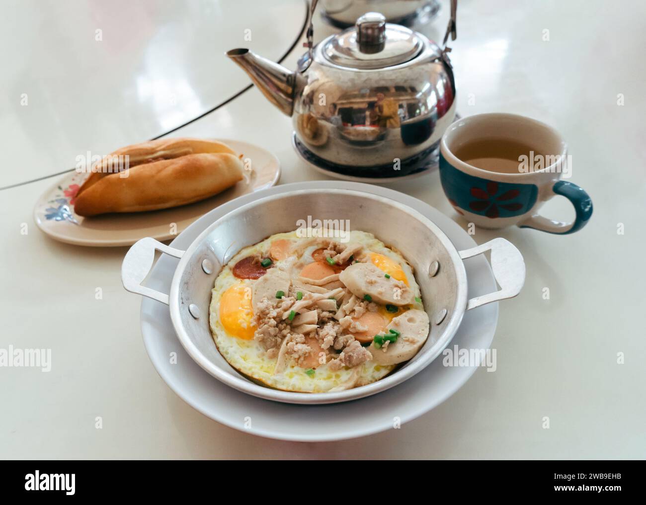 Spiegeleier in einer kleinen Pfanne mit Schweinefleisch und Würstchen, serviert auf dem Tisch mit Banh mi Schweinefleisch und Gurken vietnamesisches Sandwich und chinesischer Teekanne. Flaches Futter Stockfoto