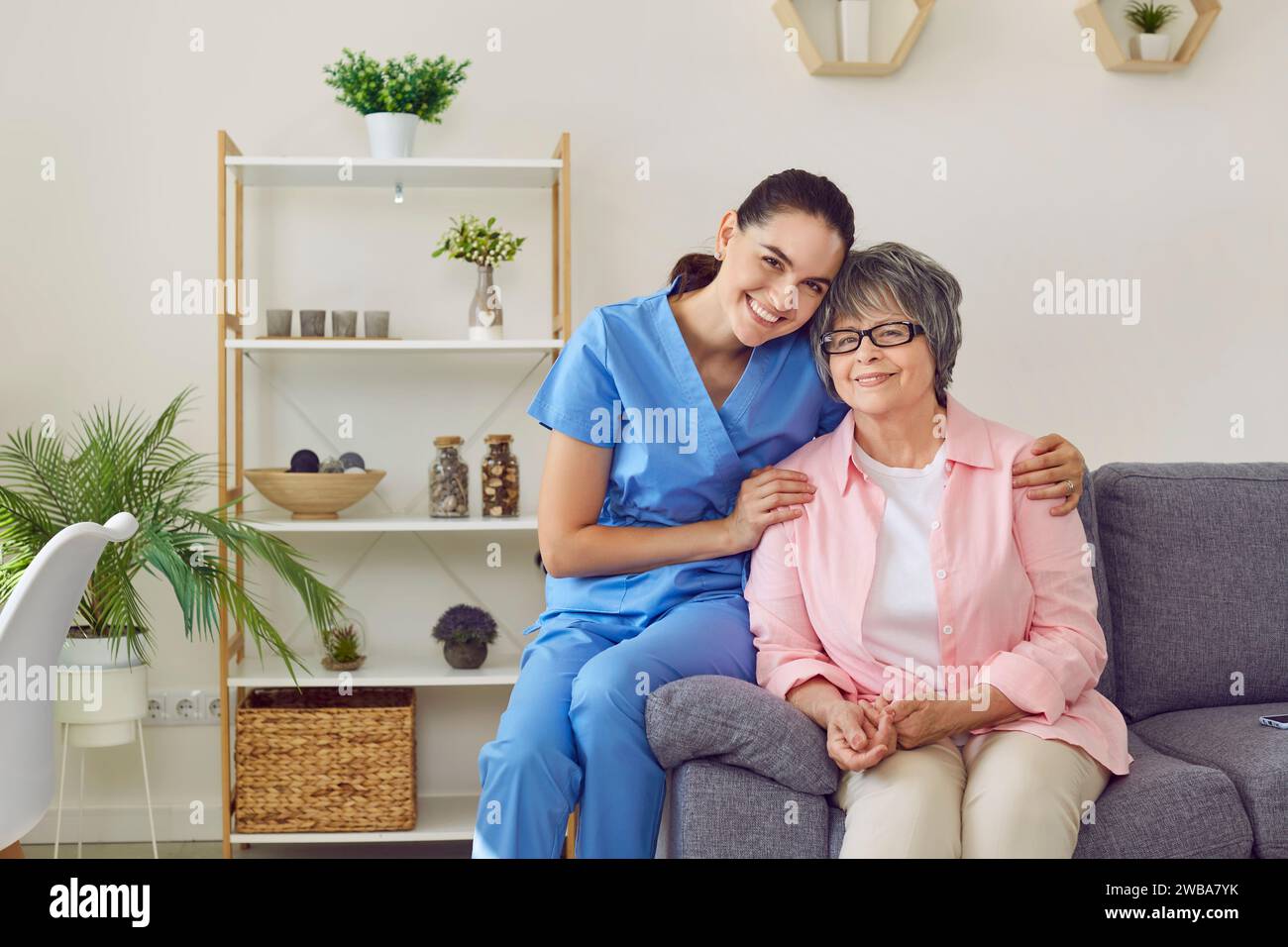 Schöne, junge, glückliche, lächelnde Krankenschwester umarmt freundlich ihre geduldige, gepflegte, hübsche grauhaarige Dame. Stockfoto