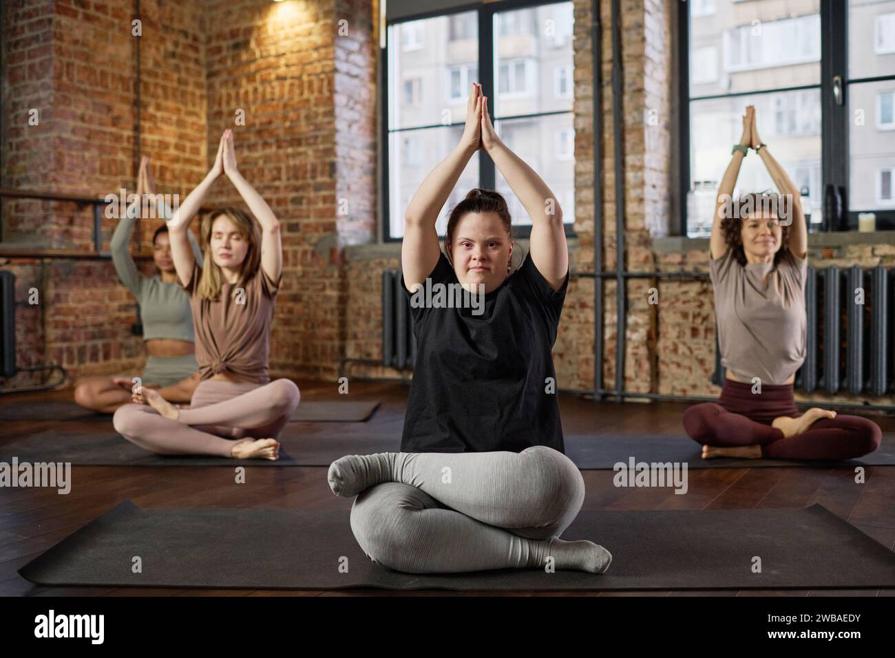 Junge aktive Frau mit Behinderung, die Beine gekreuzt und die Hände über dem Kopf zusammengelegt hält und während des Yoga-Trainings in die Kamera blickt Stockfoto