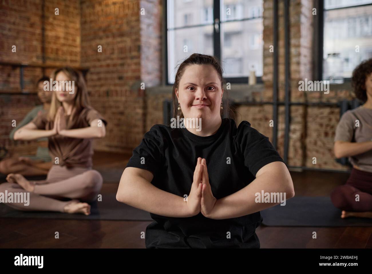 Aktives Mädchen mit Behinderung, das ihre Hände gegen die Brust legt und während des Yoga-Trainings mit anderen Frauen in die Kamera schaut Stockfoto
