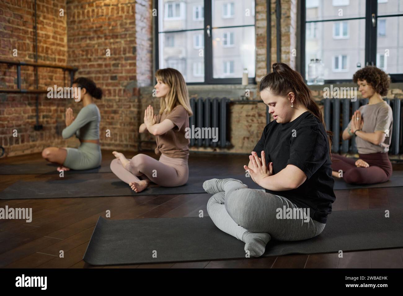 Weibliche Fitnesstrainerin mit Behinderung hält die Hände gegen die Brust, während sie auf dem Boden sitzt und Yoga im Fitnessstudio übt Stockfoto