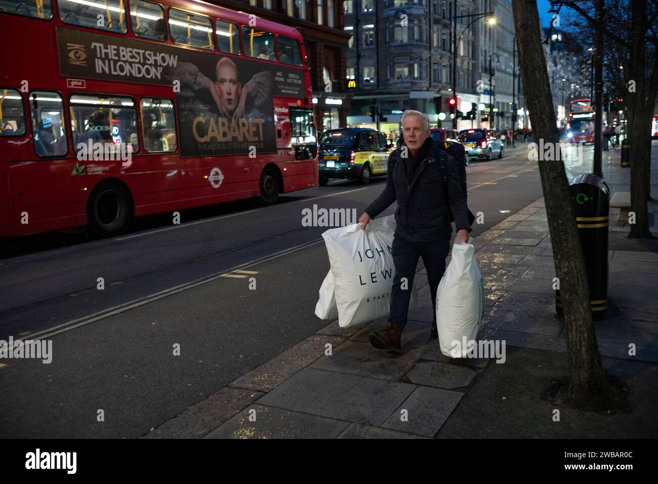 Ein Mann mit John Lewis Taschen trotzt dem kalten Winter bei eisigen Schneeregen und Regenschauern auf der Oxford Street, Londons geschäftigster High Street. Stockfoto