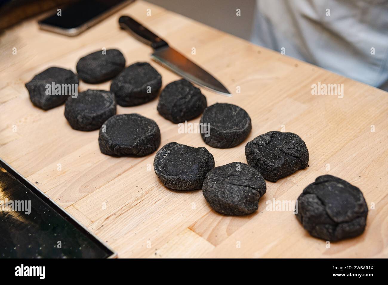Der Teig mit Tintenfischtinte wird auf einem Holzschneidebrett neben einem Kochmesser zu Brötchen geformt, was die kulinarische Zubereitung anzeigt. Stockfoto