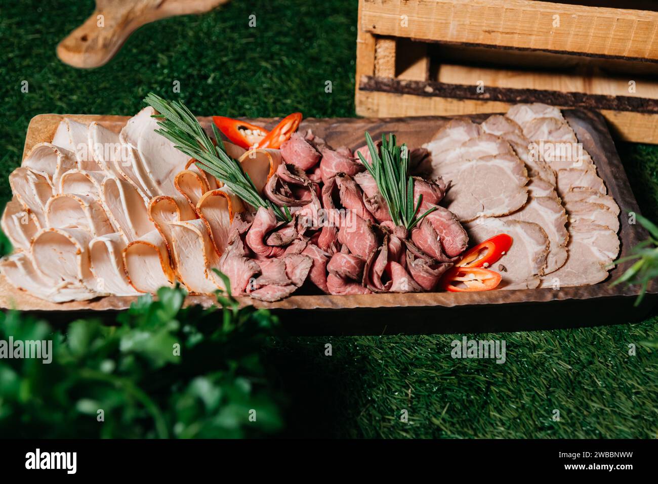 Ein rustikales hölzernes Servierbrett mit dünn geschnittenem Roastbeef und truthahn, garniert mit frischem Rosmarin und Chili-Pfeffer, präsentiert auf Kunstrasen. Stockfoto