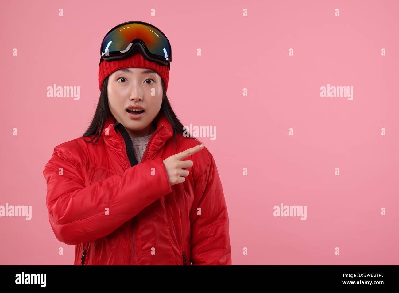 Überraschte Frau in Wintersportbekleidung, die auf etwas auf rosa Hintergrund zeigt. Leerzeichen für Text Stockfoto