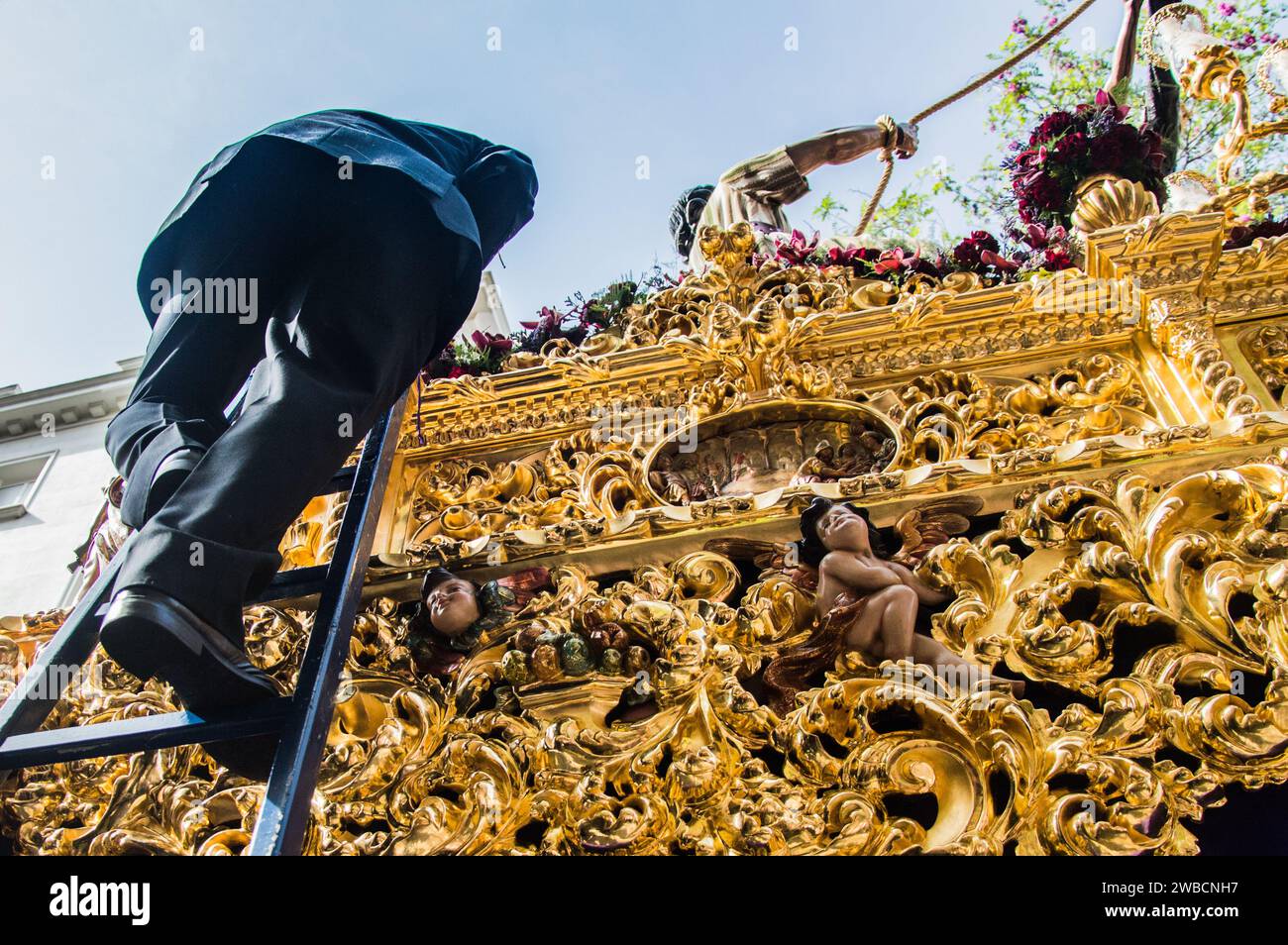 Der Gläubige steigt eine Leiter auf dem kunstvoll verzierten Festzug in Sevilla hinauf, der mit aufwändig geschnitzten Holzblättern und glitzerndem Blattgold geschmückt ist. Stockfoto