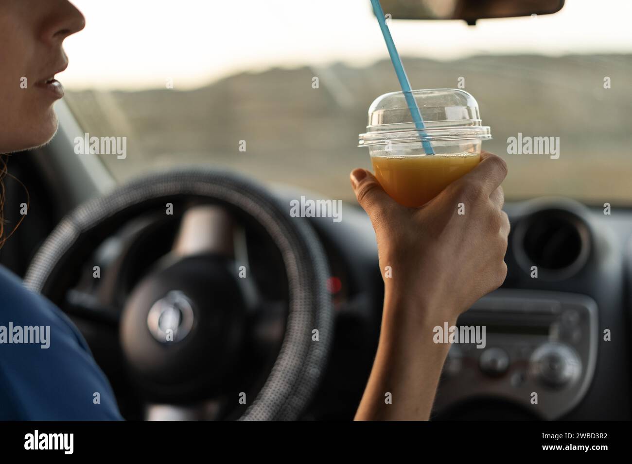 Eine Fahrerin trinkt kalten Saft aus einem Plastikglas in ihrem Auto, durstig hinter dem Steuer, hielt an, um sich auszuruhen. Stockfoto