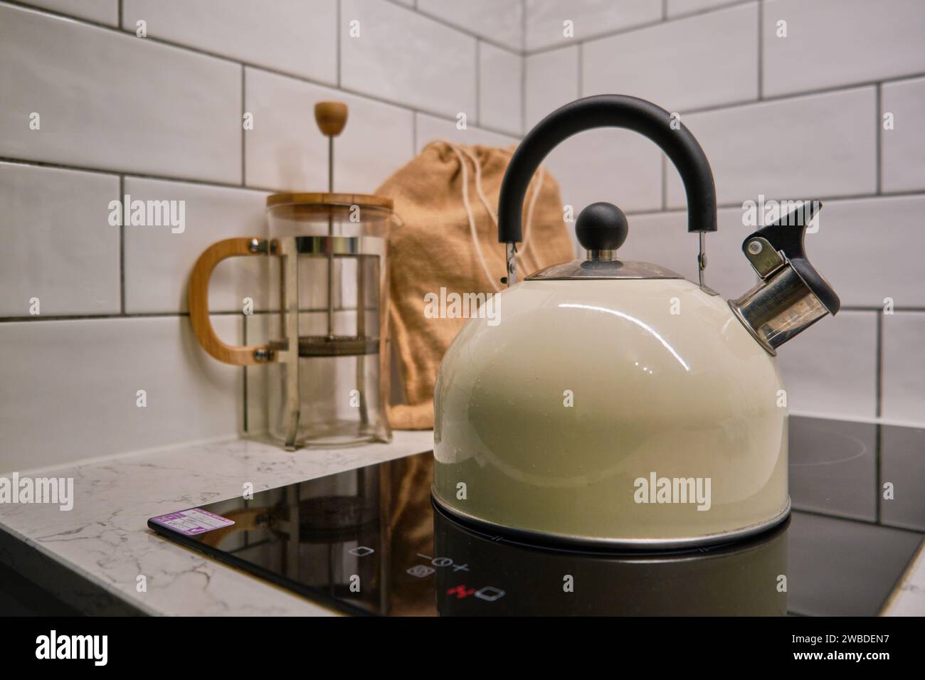 Creme, Edelstahl Pfeifkessel auf einem schwarzen Induktionskochfeld, hessische Tasche mit Kaffeebohnen, französische Presse Cafetier und weiß geflieste Küche Hintergrund. Stockfoto