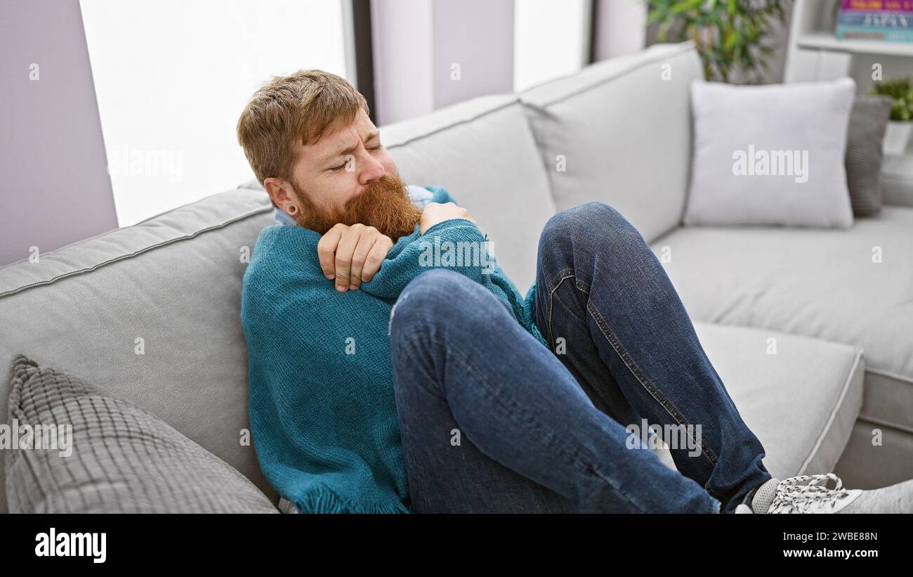Die Geschichte des kalten Winters, der hübsche junge rothaarige Mann, der friert, findet gemütlichen Komfort in einer warmen Decke, sitzt auf dem Sofa und kämpft zuhause gegen die Kälte. Stockfoto Die Geschichte des kalten Winters, der hübsche junge rothaarige Mann, der friert, findet gemütlichen Komfort in einer warmen Decke, sitzt auf dem Sofa und kämpft zuhause gegen die Kälte. Stockfoto