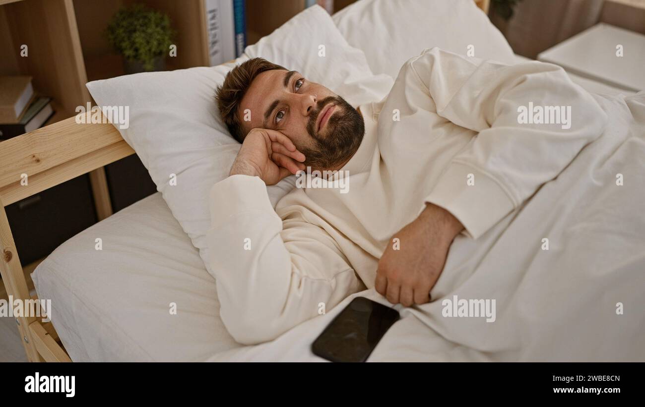 Nachdenklicher junger hispanischer Mann, der in seinem Schlafzimmer aufwacht, ernsthaft in sein Handy vertieft, bequem auf seinem Bett liegend, in seiner Morgendecke eingekocht Stockfoto
