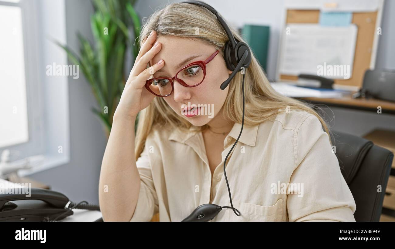 Eine gestresste junge blonde Frau mit Brille und Headset in einem Büro. Stockfoto