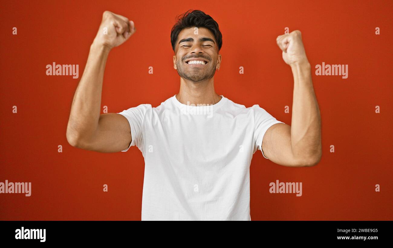 Hübscher junger hispanischer Mann, der triumphierend vor einem leuchtend roten Hintergrund feiert Stockfoto
