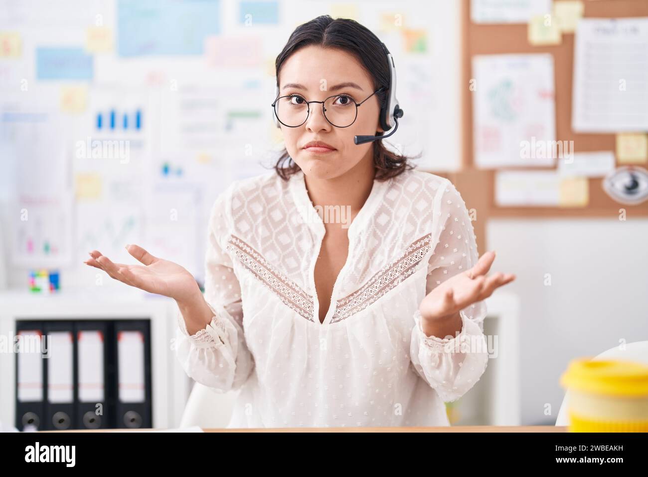 Hispanische junge Frau, die im Büro arbeitet, mit Headset und Brille, laut schreiend und mit Hand auf Mund. Kommunikationskonzept. Stockfoto