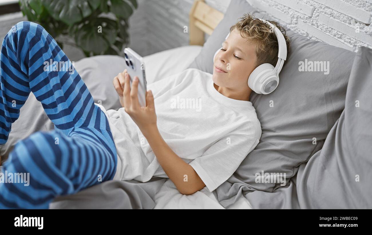 Entzückender blonder Junge, in einer Decke gekuschelt, entspannt auf einem Bett in einem gemütlichen Zimmer, vertieft und konzentriert sich ernsthaft auf ein Video auf seinem Smartpho Stockfoto