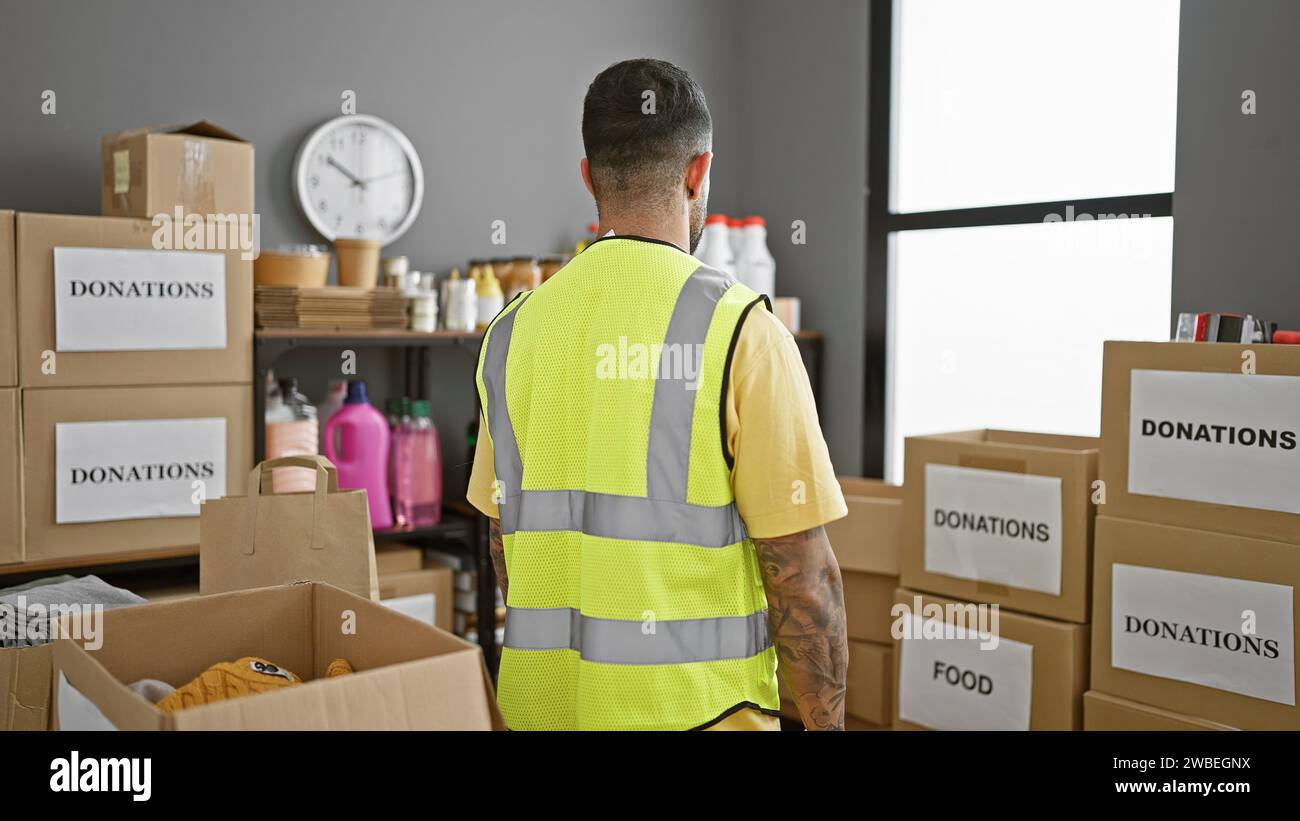 Hispanischer Mann in reflektierender Weste, der Spenden in einem Lagerhaus mit Boxen mit der Aufschrift „Essen“ und „Spenden“ überwacht. Stockfoto