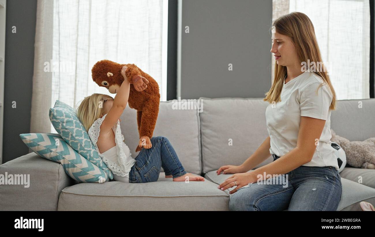 Kaukasische Mutter, die sich zu Hause auf dem Sofa entspannt, verspielt in der Welt des Scheinglaubens mit ihrer kleinen Tochter und einer Affenpuppe Stockfoto