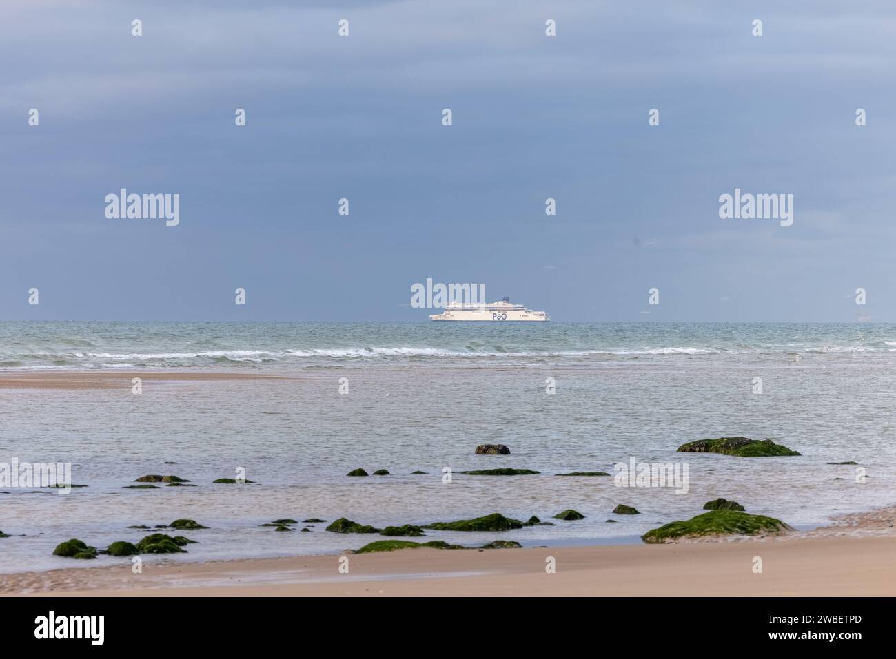 In dieser ruhigen Küstenlandschaft wird ein weit entferntes Passagierschiff gefangen, das über den Horizont segelt, eine scharf weiße Präsenz vor dem Hintergrund eines gedämpften Meeres und Himmels. Im Vordergrund befindet sich eine friedliche Küste mit verstreuten Felsen, die mit Algen bedeckt sind, Symbole für das ruhige Leben unter den Wellen. Das Foto vermittelt die Weite des Ozeans und die Reise des Schiffes, während es dieses weitläufige Blau durchquert, eine subtile Erinnerung an die unaufhörliche Bewegung und Verbindung über das Wasser. Seefahrt: Schiff am Horizont. Hochwertige Fotos Stockfoto