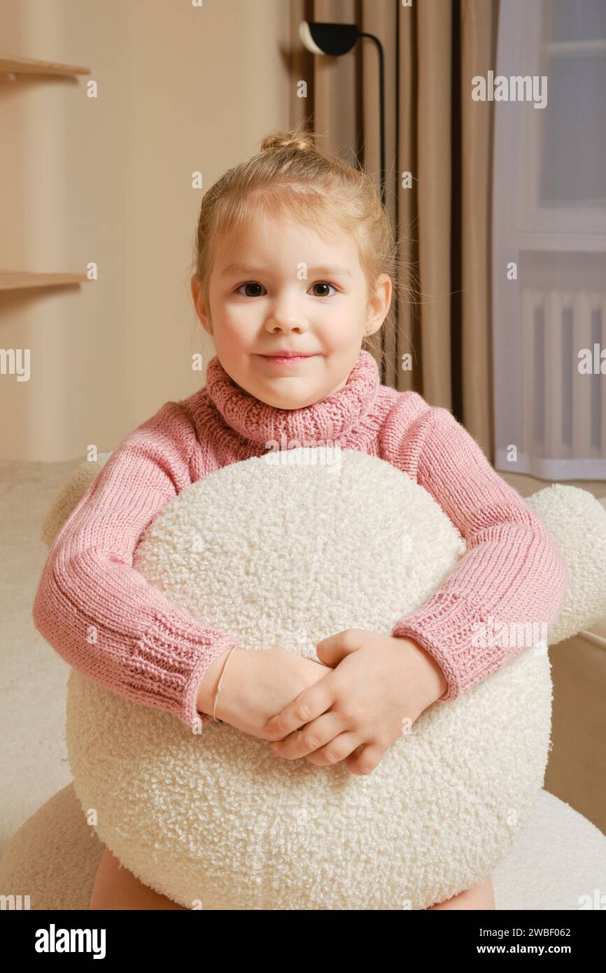 In einem gemütlichen Zimmer umschließt das kleine Mädchen in rosa Pullover ein großes weißes, gefüttertes Dekokissen Stockfoto