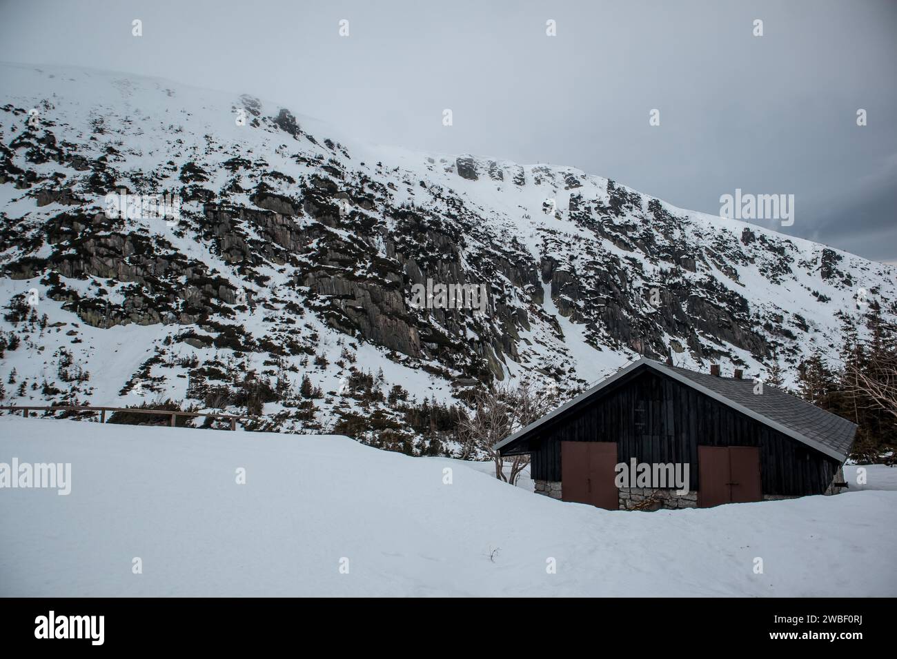 Eine einsame Hütte befindet sich unter einer zerklüfteten, schneebedeckten Berglandschaft unter einem bewölkten Himmel. Polen, Karpacz, Karkonosze Stockfoto