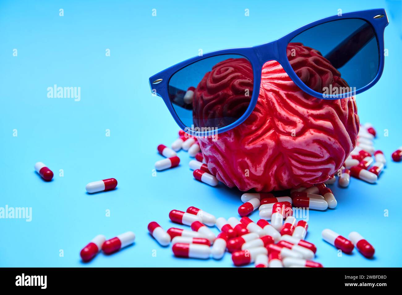 Rotes Gummigehirn mit Sonnenbrille umgeben von weißen und roten Kapseln auf blauer Oberfläche. Migräne- und Kopfschmerz-Konzept. Stockfoto