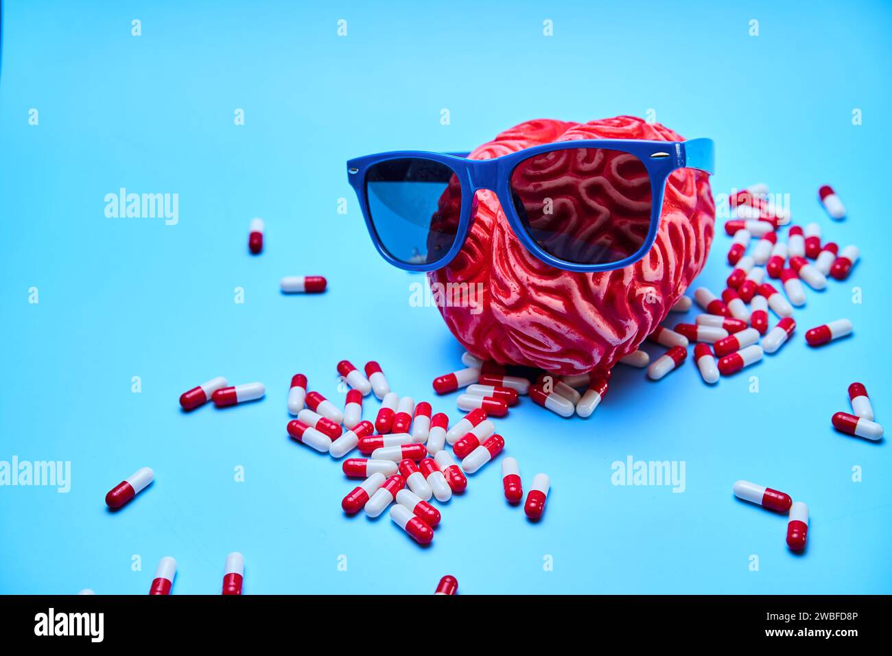 Rotes Gehirn mit Sonnenbrille und ein Haufen Pillen drumherum, auf einer blauen Oberfläche. Stockfoto