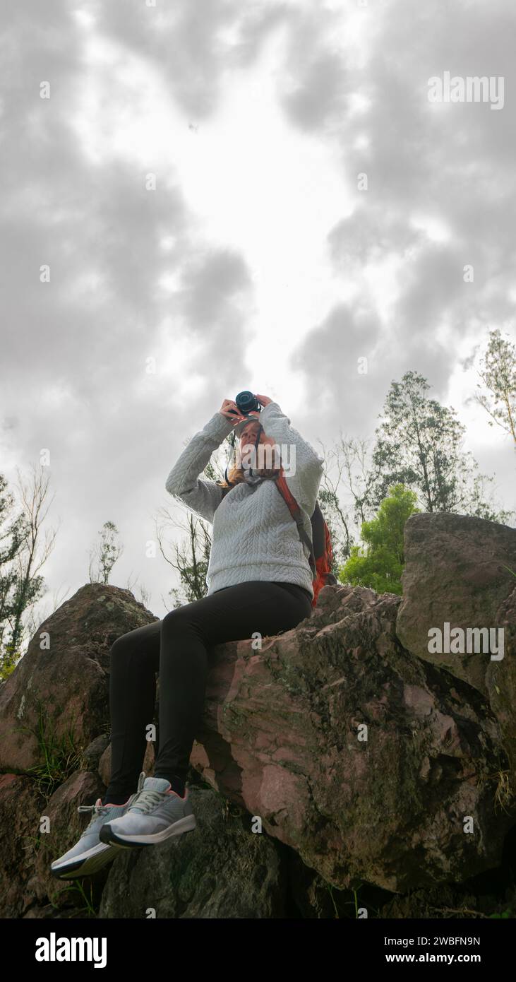 Schöne junge lateinamerikanische Frau, die auf Steinen auf einem Berg sitzt und an einem bewölkten Tag ein Foto mit ihrer kleinen Kamera macht Stockfoto