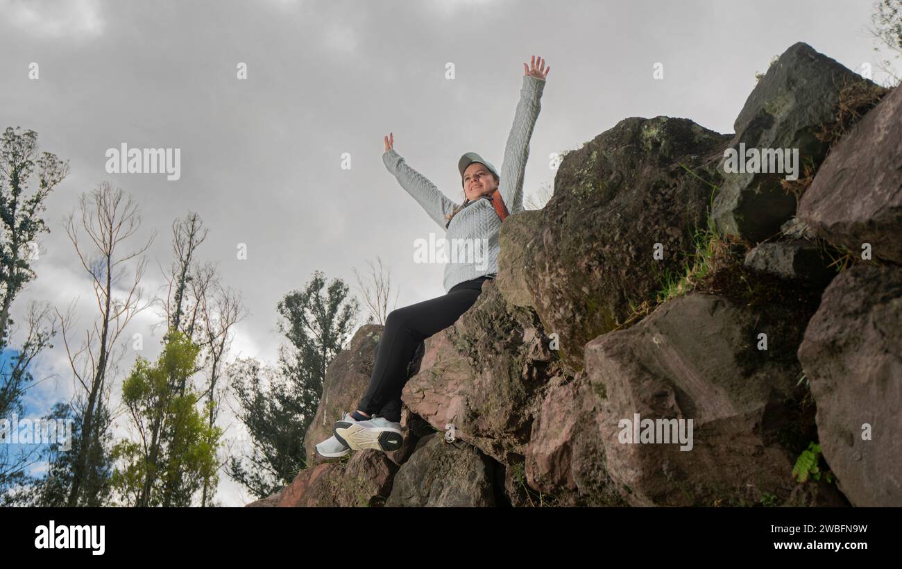 Schöne junge lateinamerikanische Frau, die auf Steinen auf einem Berg sitzt, mit erhobenen Armen an einem bewölkten Tag Stockfoto