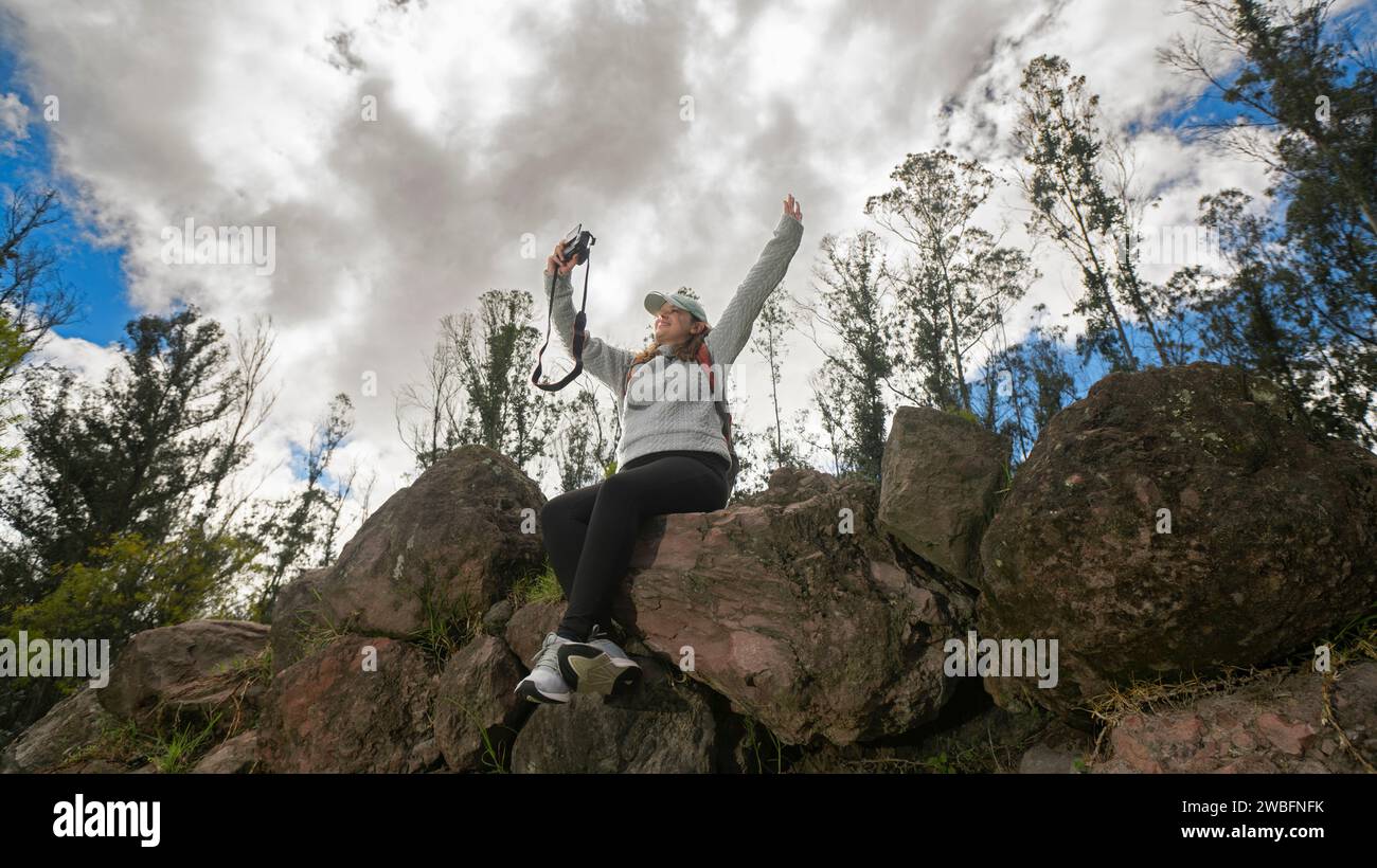 Schöne junge lateinamerikanische Frau, die auf Steinen auf einem Berg sitzt und an einem bewölkten Tag ein Selfie mit ihrer kleinen Kamera macht Stockfoto