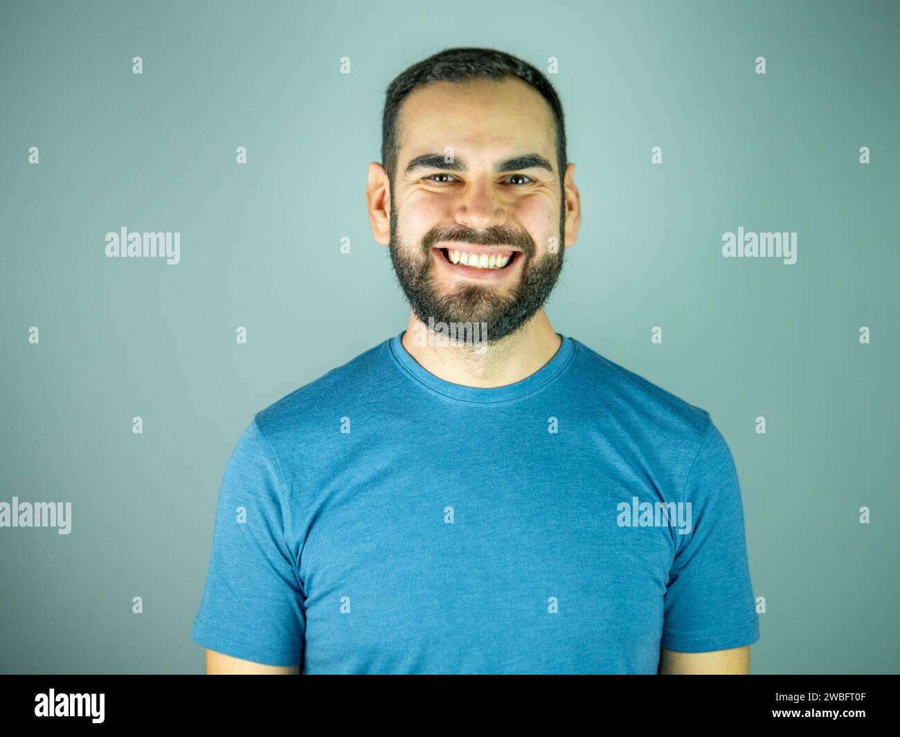 Junger bärtiger Mann mit blauem T-Shirt, der lächelt und in die Kamera blickt, in einem grauen Brackground Stockfoto