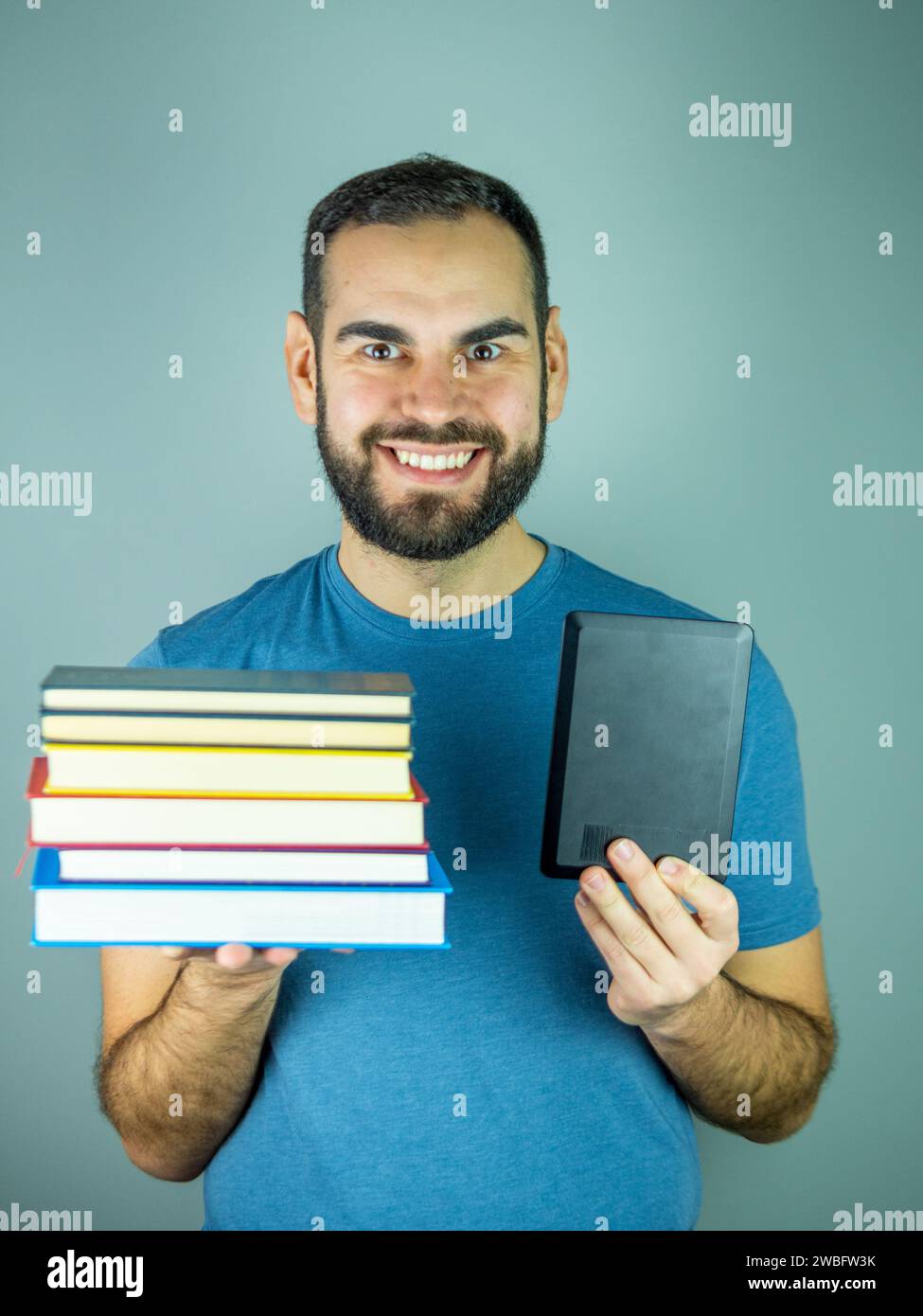 Junger bärtiger Mann mit einem Stapel Bücher in der einen Hand und einem elektronischen Buch in der anderen Stockfoto