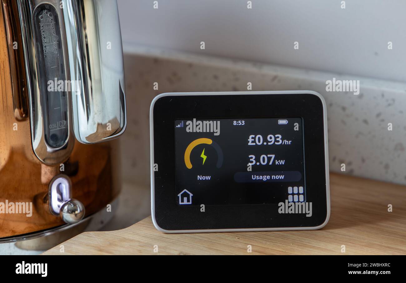 Intelligenter Energiemonitor mit Anzeige des Stromverbrauchs und der Kosten pro Stunde auf einer Küchenarbeitsplatte neben einem Wasserkocher Stockfoto