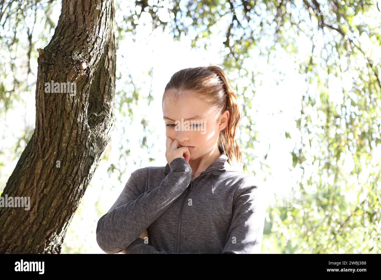 Teenager-Mädchen mit grauem Oberteil unter grünen Blättern, Sonnenschein und Baumstamm, ängstlich und besorgt mit den Augen nach unten Ökoangst Stockfoto