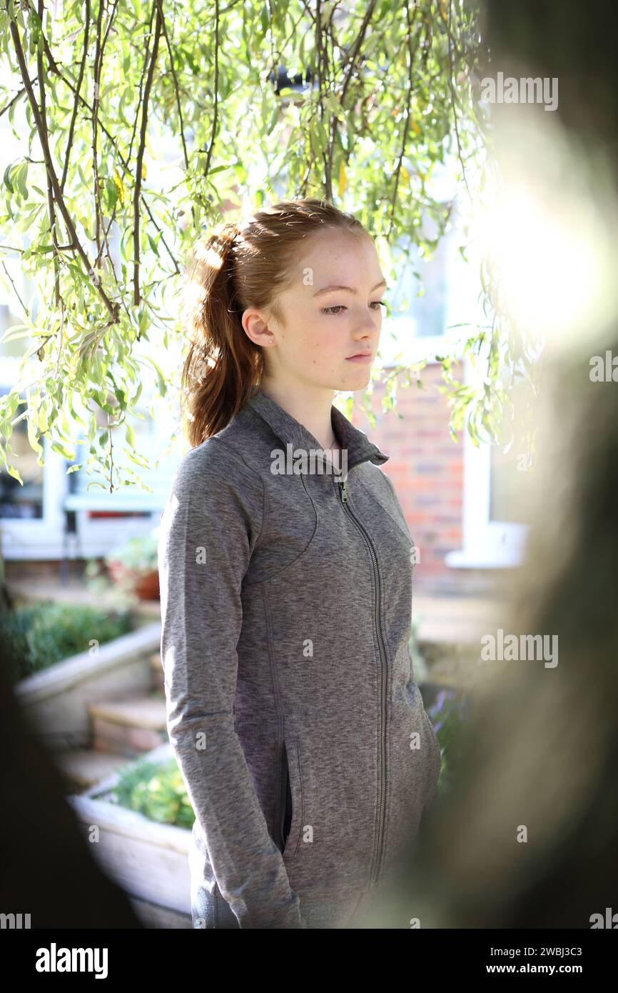 Teenager-Mädchen mit grauem Oberteil unter grünen Blättern, hinterleuchtet von Sonnenschein und Baumstamm-Rahmen-Porträt Stockfoto