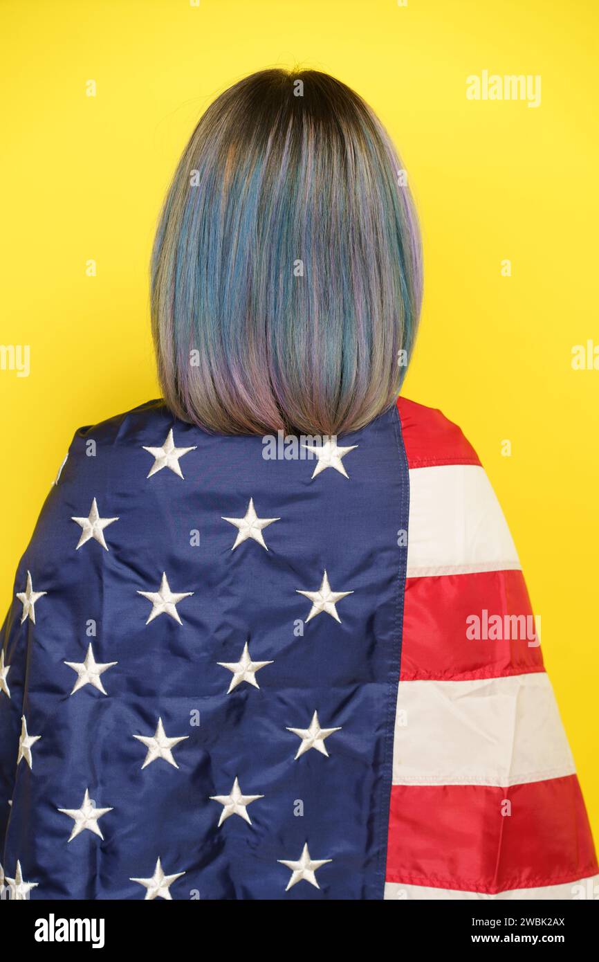 Patriotischer Stolz Und Freiheitsgeist Der Vereinigten Staaten Und Ihr Kultureller Reichtum. Mädchen Mit Bunten Haaren Bedeckt Mit Amerikanischer Flagge, Isoliert Auf Gelbem Hintergrund, Rückansicht. Hochwertige Fotos Stockfoto