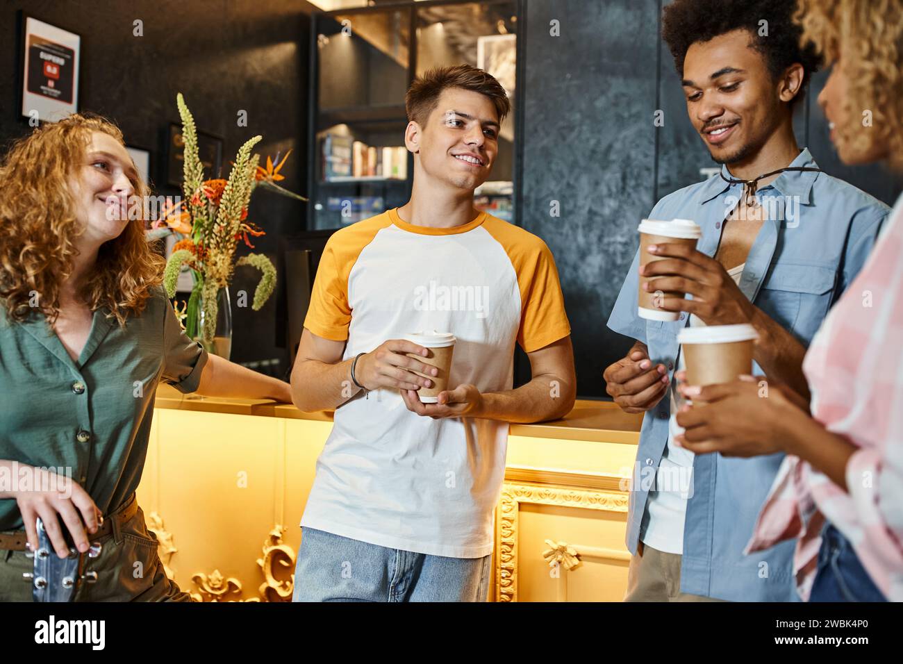 Fröhliche multiethnische Freunde mit Getränken zum Mitnehmen, die sich an der Rezeption in der Jugendherberge unterhalten, reisen Stockfoto