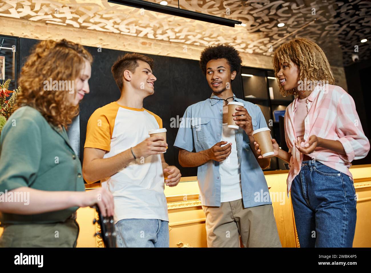 Fröhliche und stilvolle multiethnische Freunde mit Kaffee, um sich an der Rezeption im Hostel zu unterhalten Stockfoto