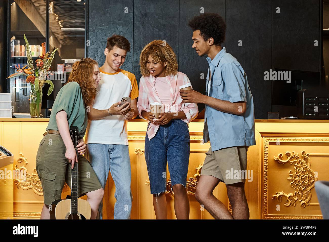 Student zeigt Smartphone multiethnischen Freunden mit Kaffee zum Mitnehmen und Gitarre in der Lobby des Hostels Stockfoto